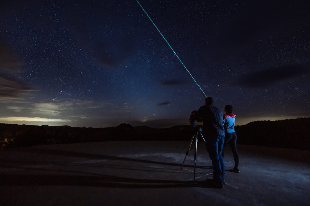 Two people on a rooftop observe the night sky, pointing a laser at the stars.